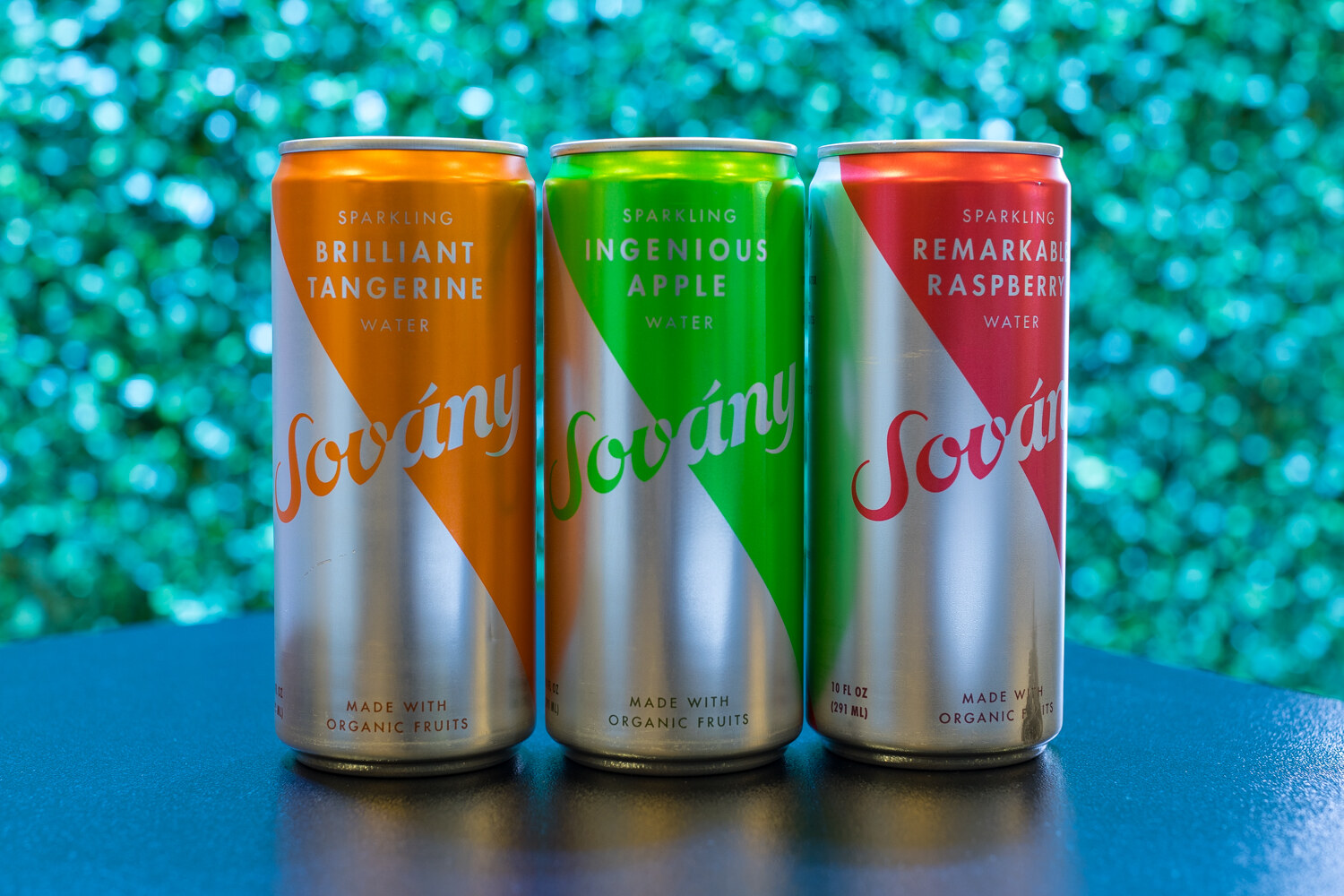 Three slim silver cans with orange, green and red swooshes labeled Sovany sparkling water, in front of a glittery teal background.