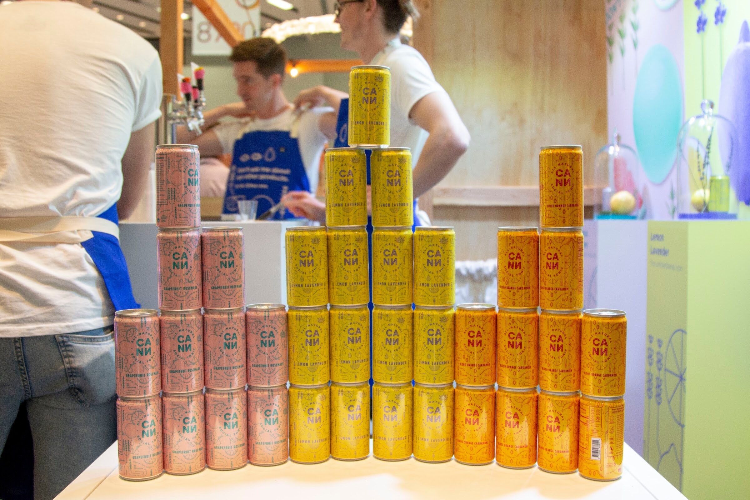 Rows of small pastel and yellow cans stacked on a white table with two blue-aproned staffers behind.