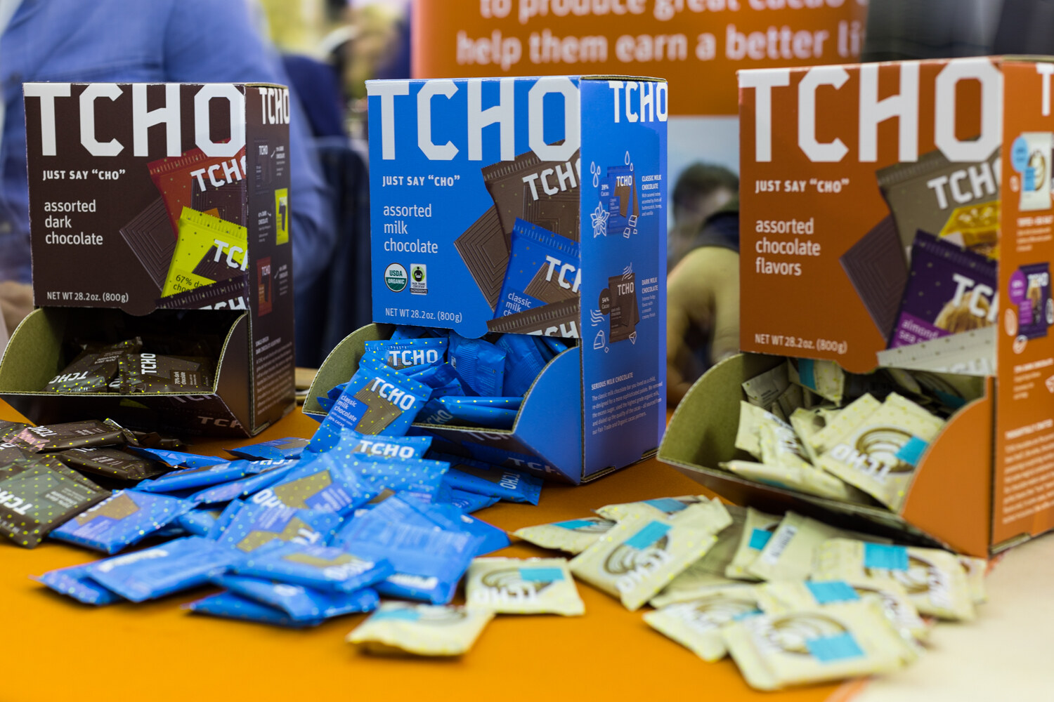 Three cardboard TCHO display boxes in brown, blue and orange with blue and cream wrapped chocolate squares scattered on an orange tabletop.