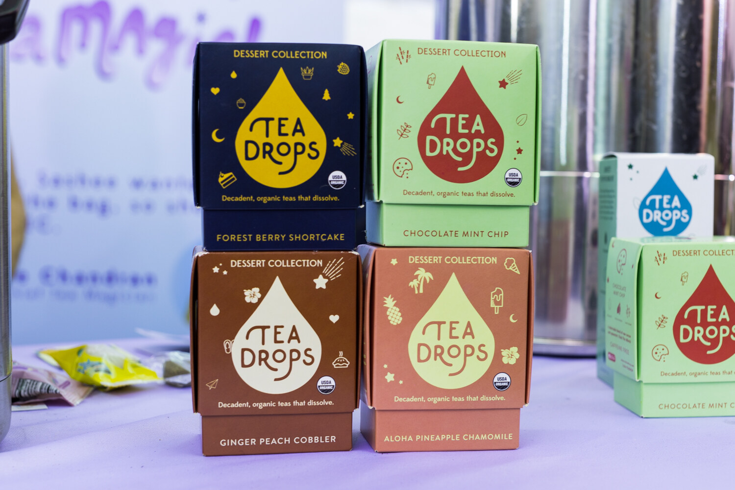 Stacked square boxes of Tea Drops in navy, mint, brown and peach with teardrop logos on a lavender tablecloth.