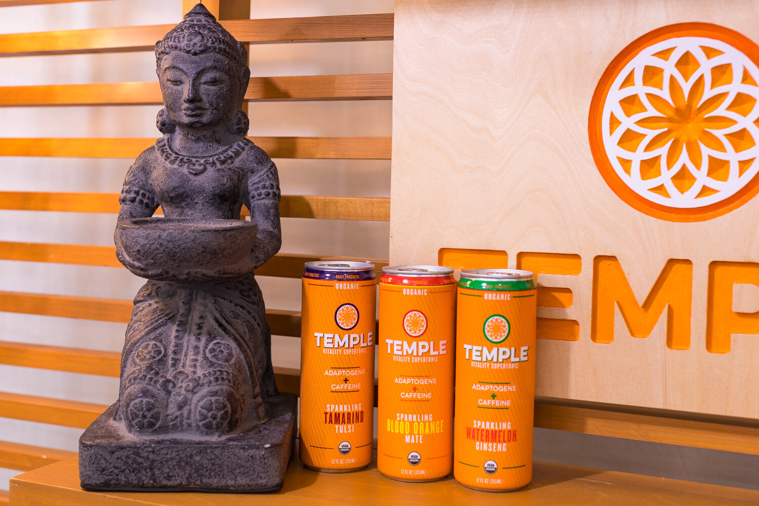 Three orange Temple cans next to a gray stone statue of a seated figure with a wooden slatted wall and a cutout Temple logo behind.