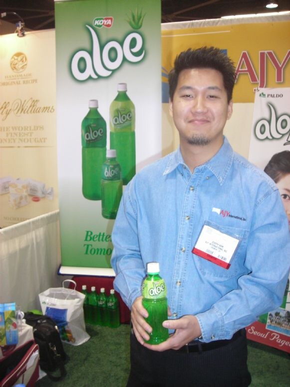 A smiling man in a blue denim shirt with an AJY International logo holds a bottle of Koya aloe drink in front of Koya and Paldo aloe banners.