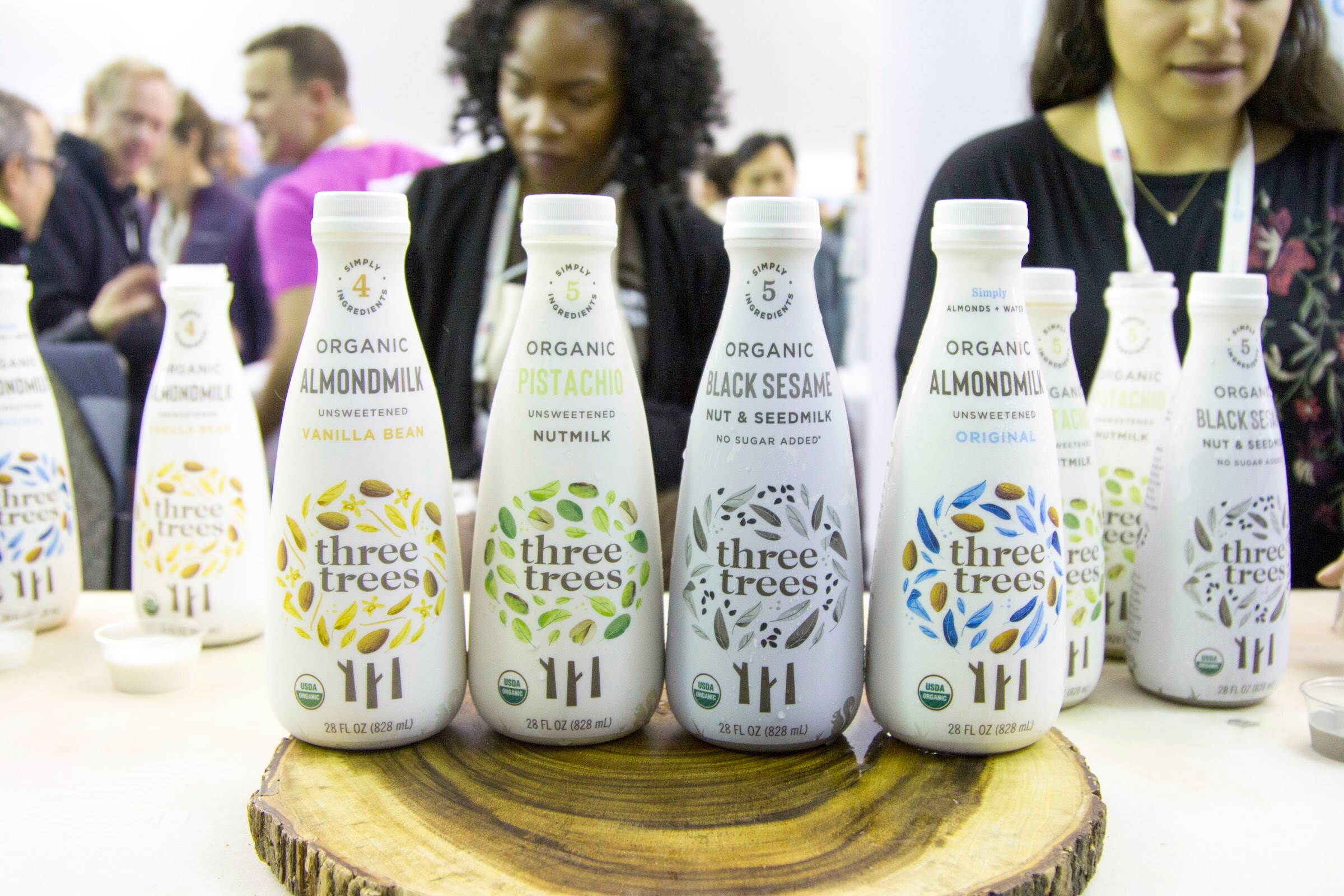 Bottles of Three Trees nut milks on a round wood slice with several attendees standing behind them.