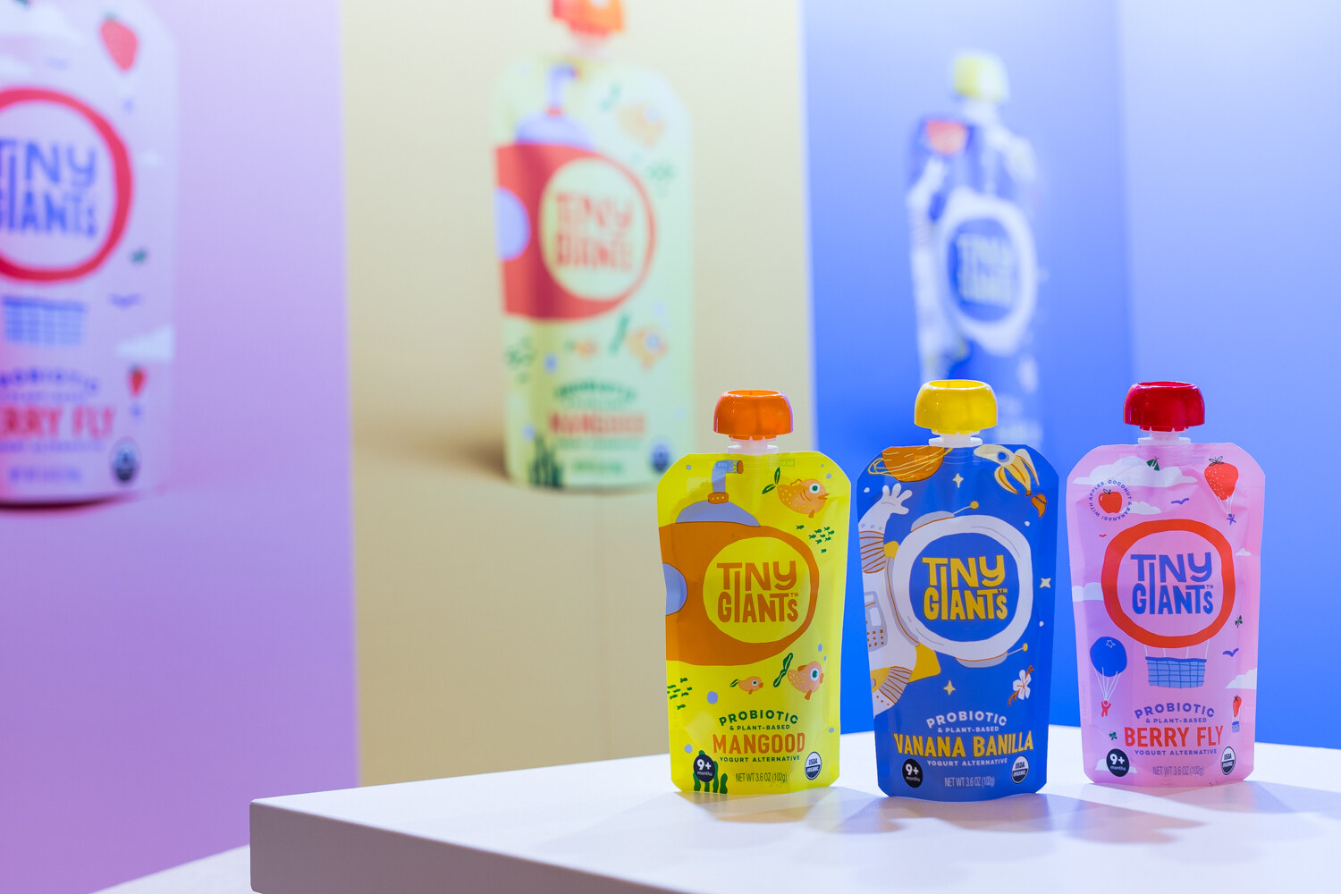 Three kids' pouches in yellow, blue and pink on a white plinth with oversized Tiny Giants pouches blurred behind.