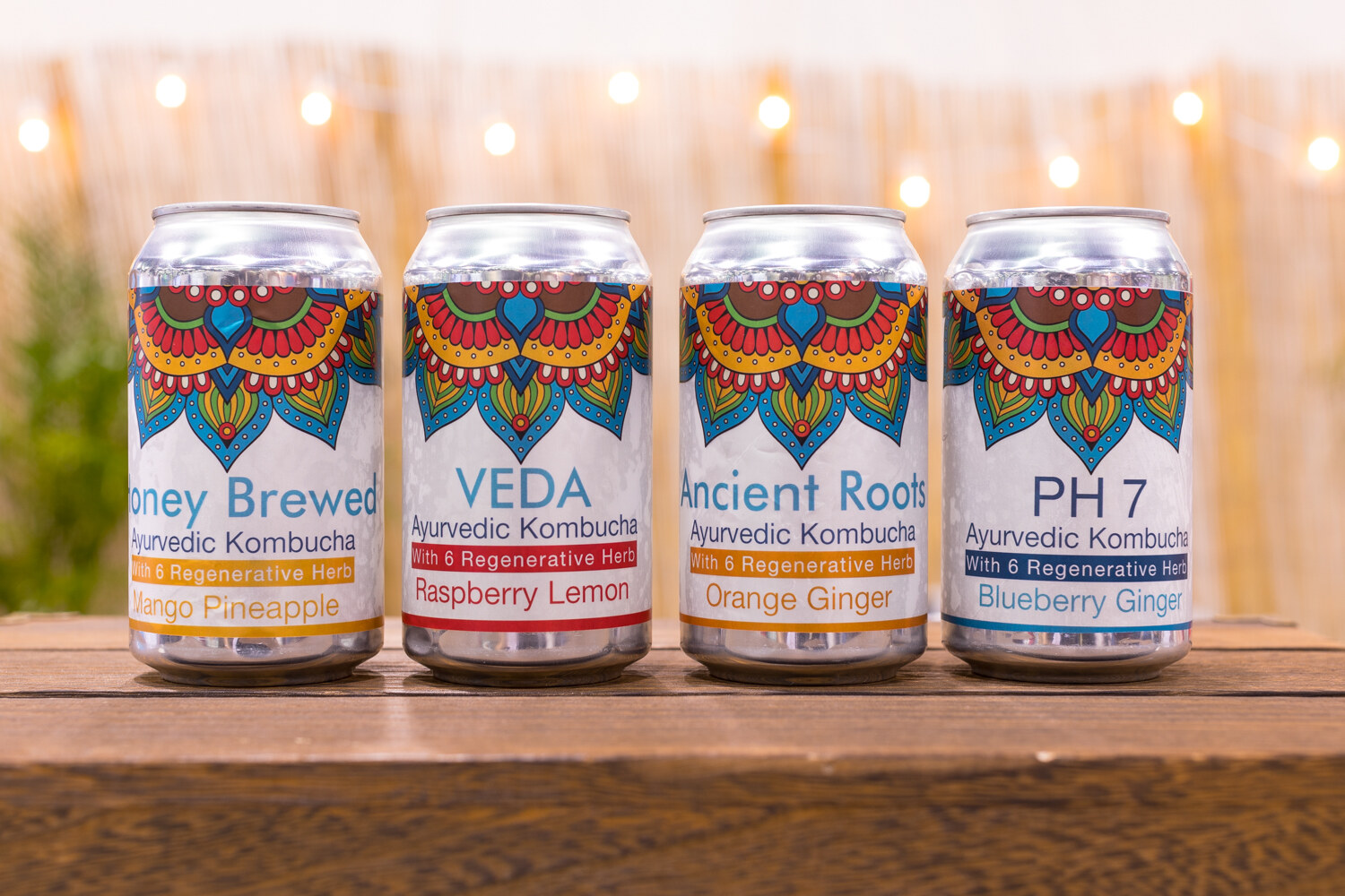 Four silver cans with mandala patterns on a wooden table with a lit bamboo-fence backdrop.