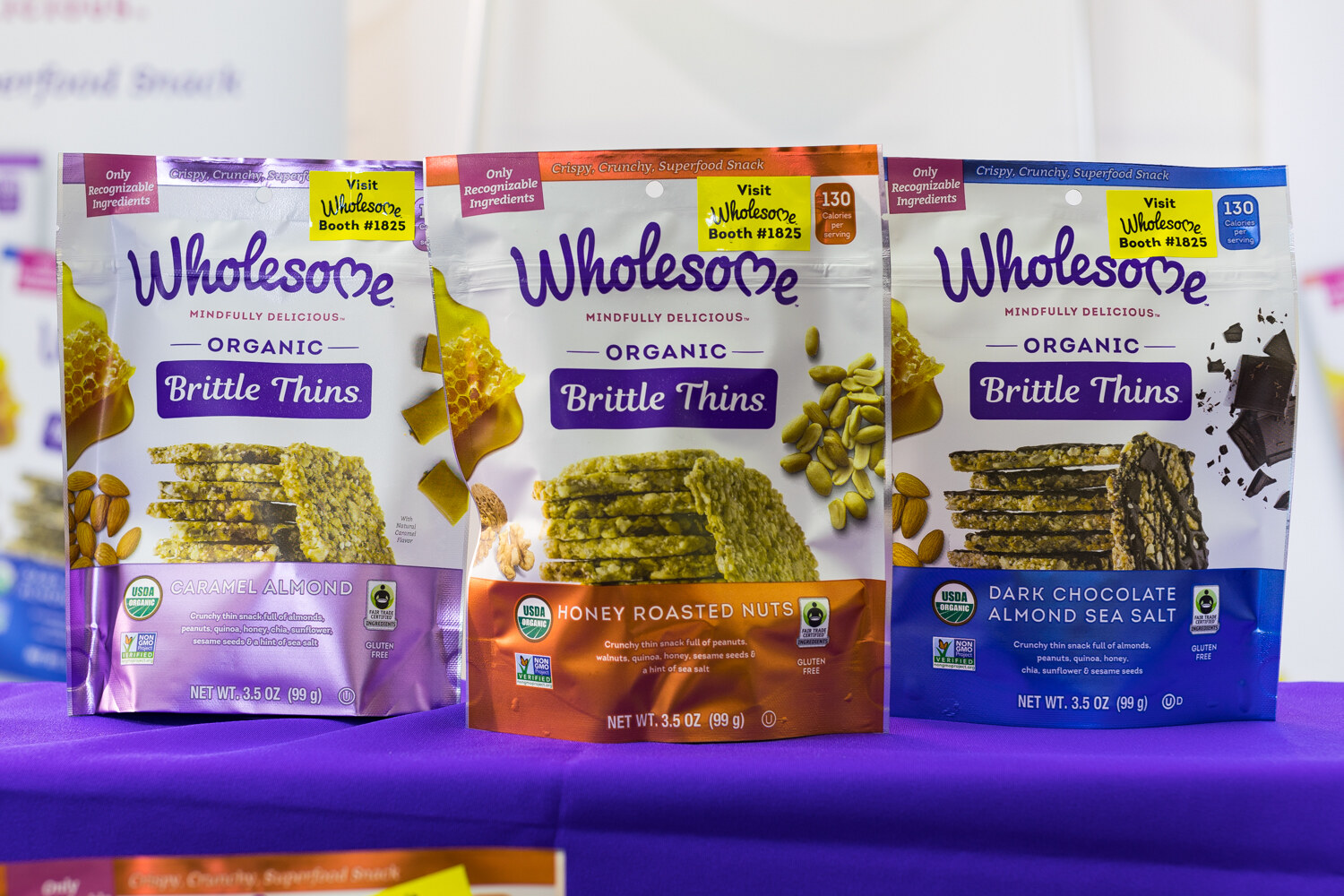 Three upright Wholesome Brittle Thins pouches in purple, orange and blue on a purple table.