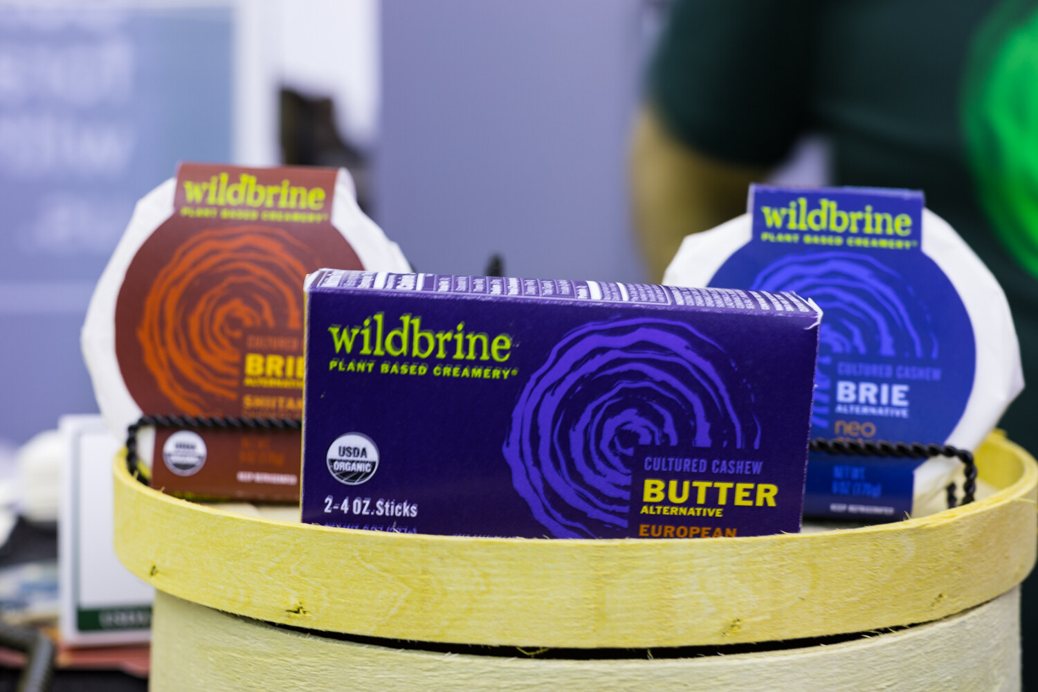 Purple Wildbrine butter alternative box in front of two round brie packages in orange and blue on a pale yellow wooden stand.