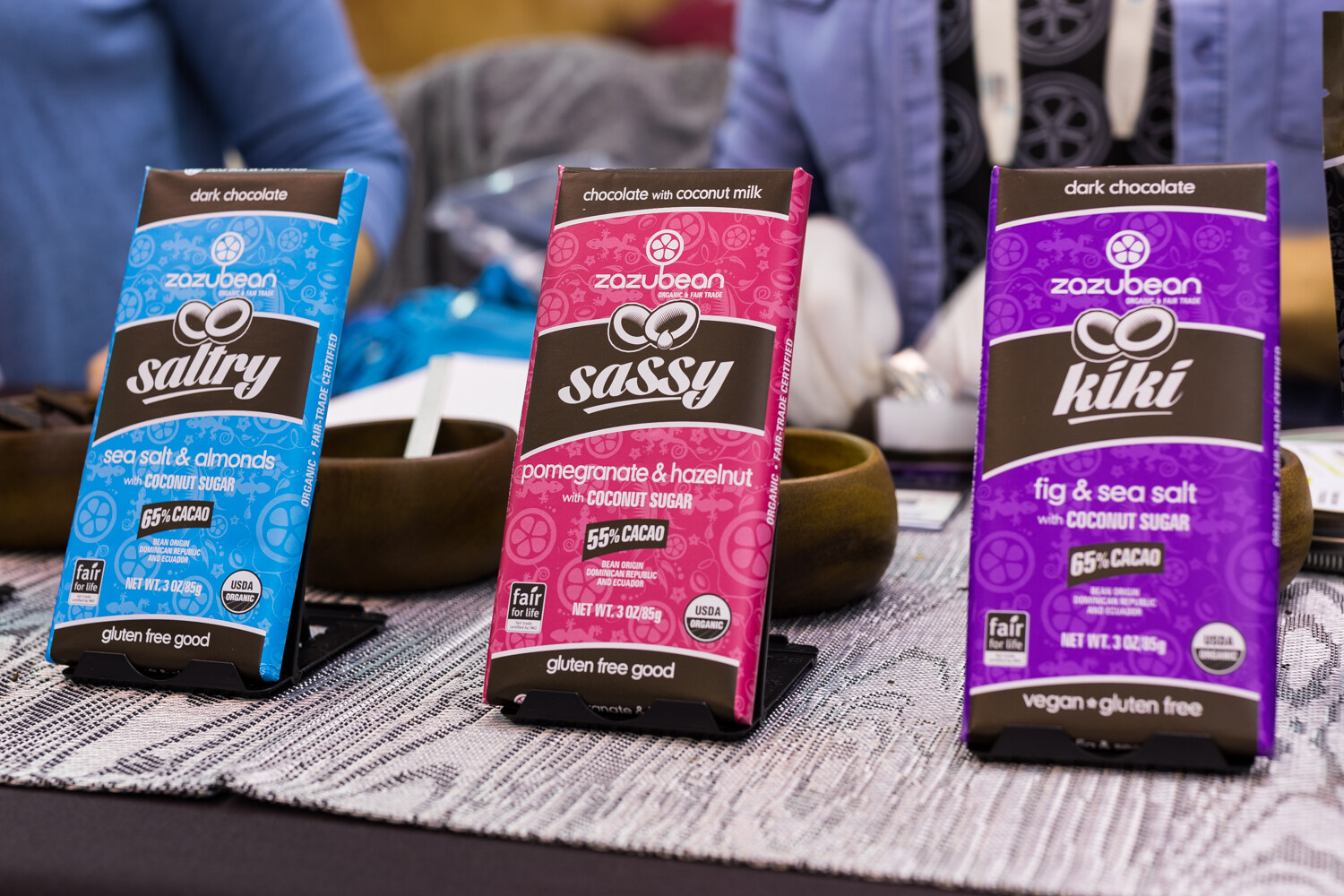 Blue, pink and purple Zazubean chocolate bars on stands with wooden bowls and a person's hands in gloves blurred behind.