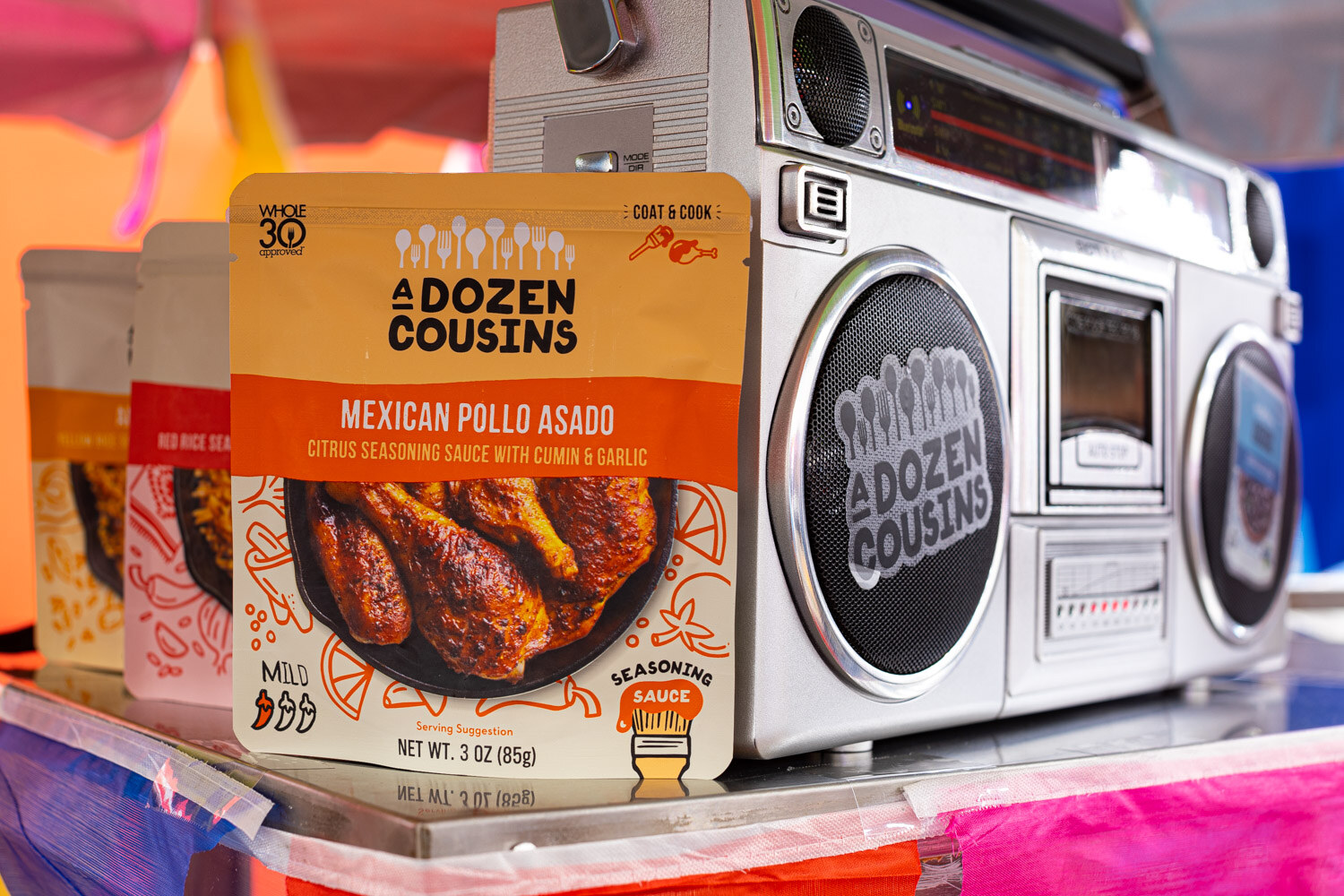 A pouch of A Dozen Cousins Mexican Pollo Asado seasoning sauce stands in front of a silver boombox