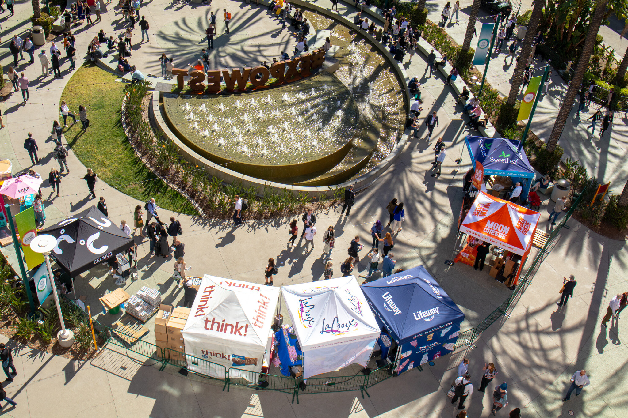 A bird's-eye view of the EXPOWEST sign on a round fountain with sampling tents for brands such as think! and LaCroix nearby