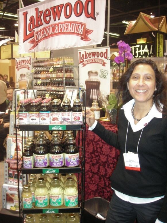 A smiling woman with dark hair stands beside a rack of Lakewood Organic juice bottles, including Pure Noni and Aloe, under Lakewood Organic & Premium banners.