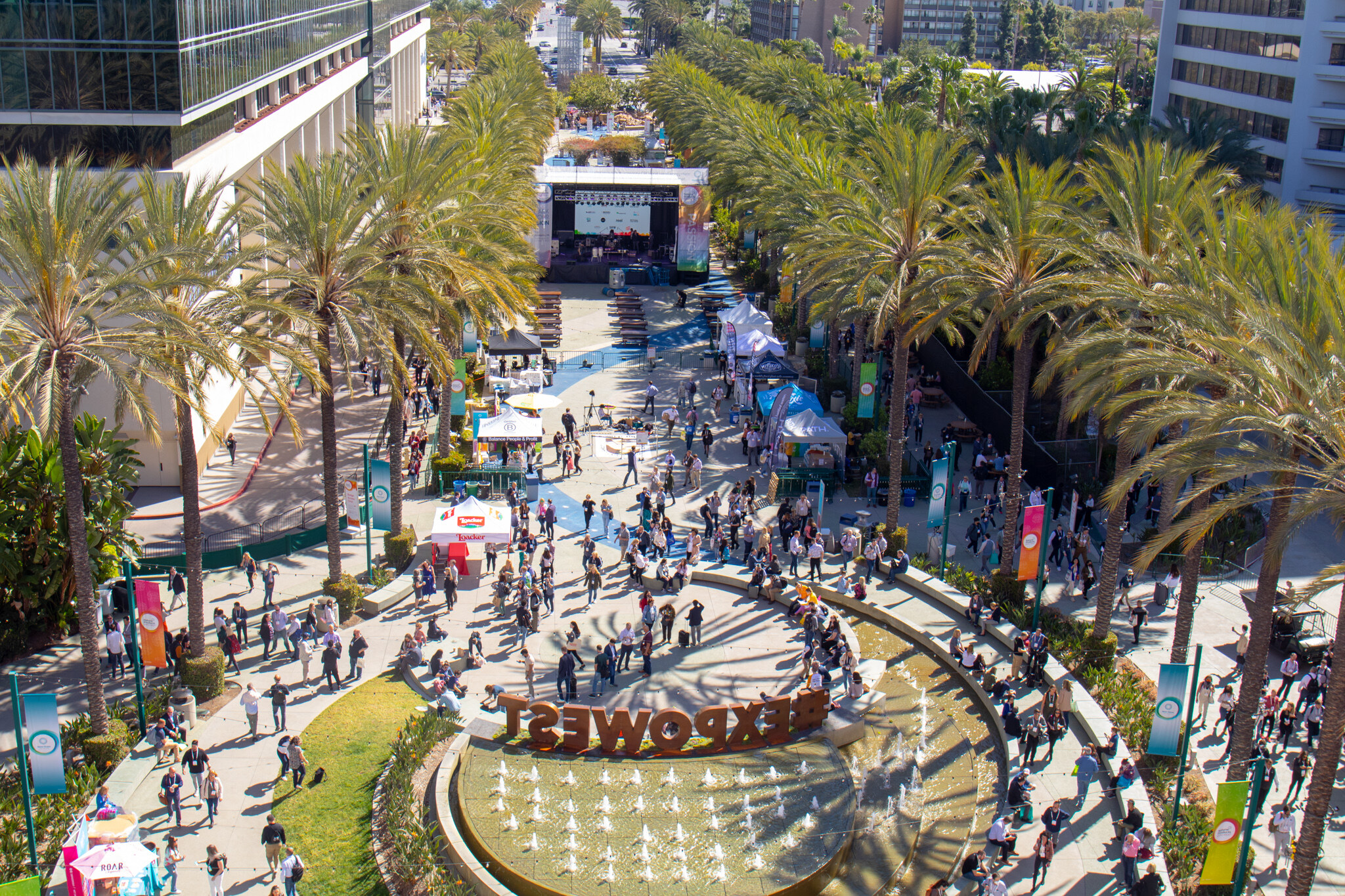 A high aerial view of the palm-lined plaza with the EXPOWEST fountain sign, outdoor stage and crowds of attendees