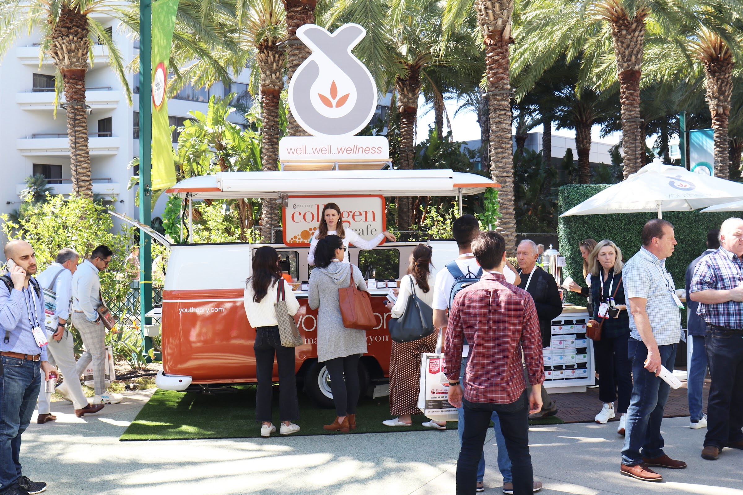 People wait at an orange and white vintage-van style cart serving a collagen drink beneath palm trees