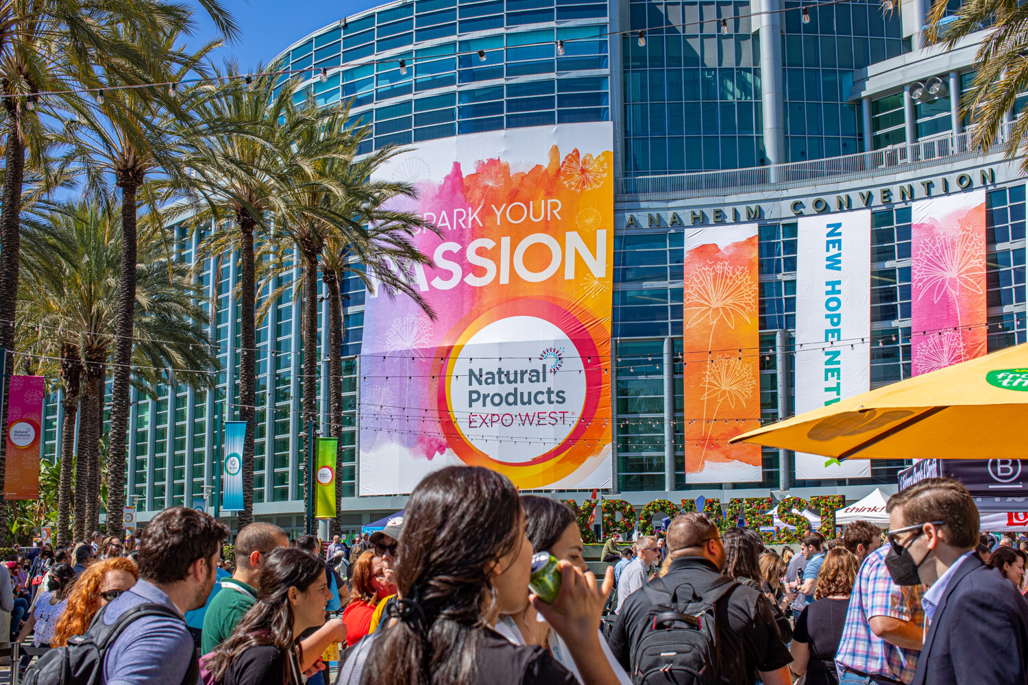 A crowd of attendees stands in the sunshine below a giant Natural Products Expo West Spark Your Passion banner on the convention center facade