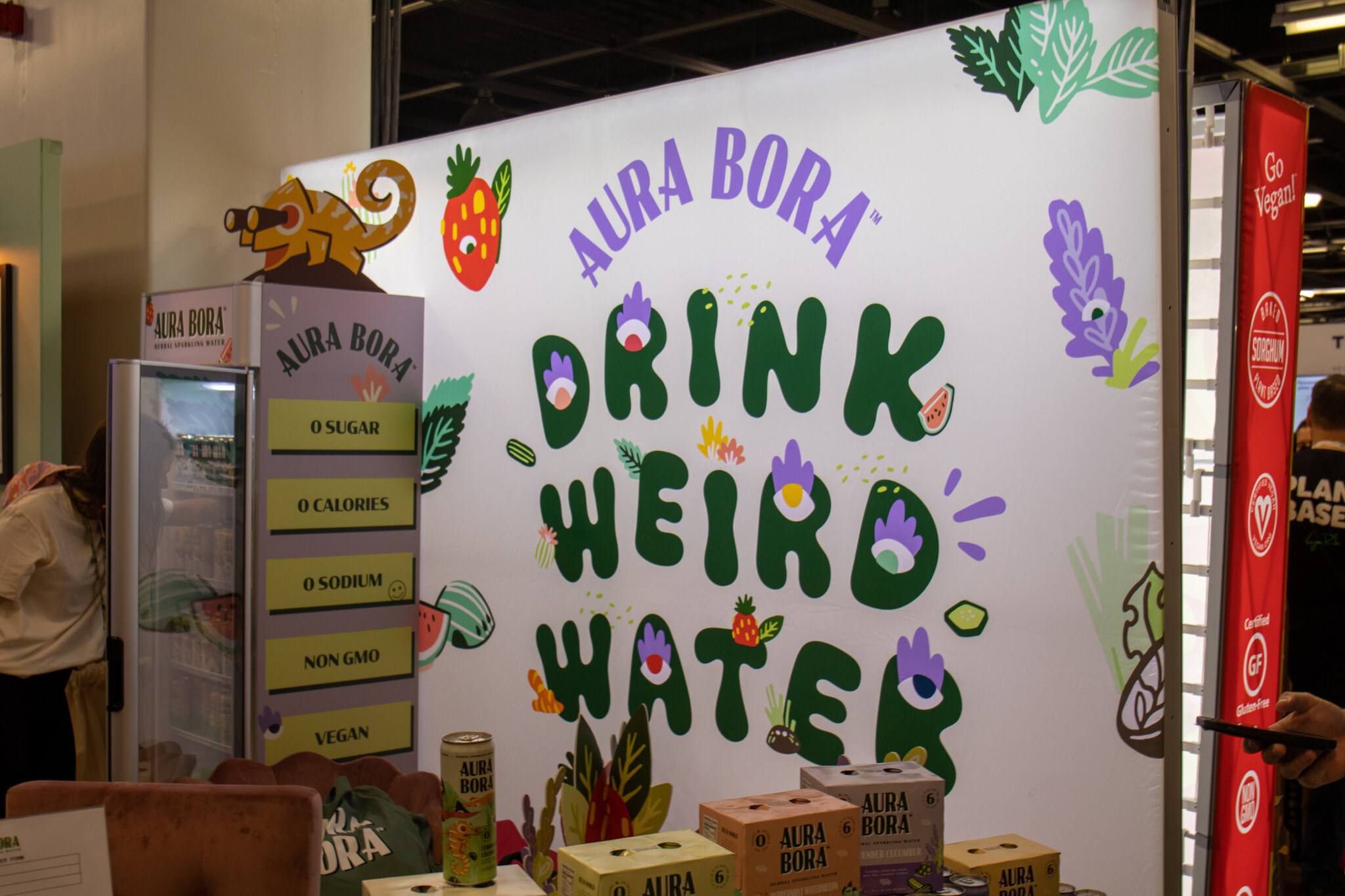 A white Aura Bora booth wall with illustrated plants and a creature reads Drink Weird Water above a cooler and boxes