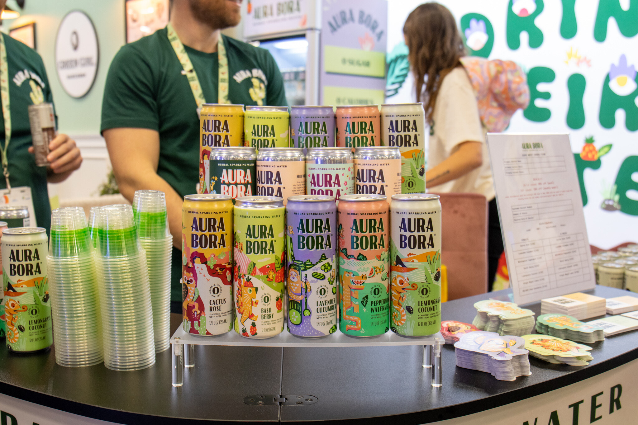Staff in green shirts stand behind tiered rows of Aura Bora herbal sparkling water cans and stacks of cups