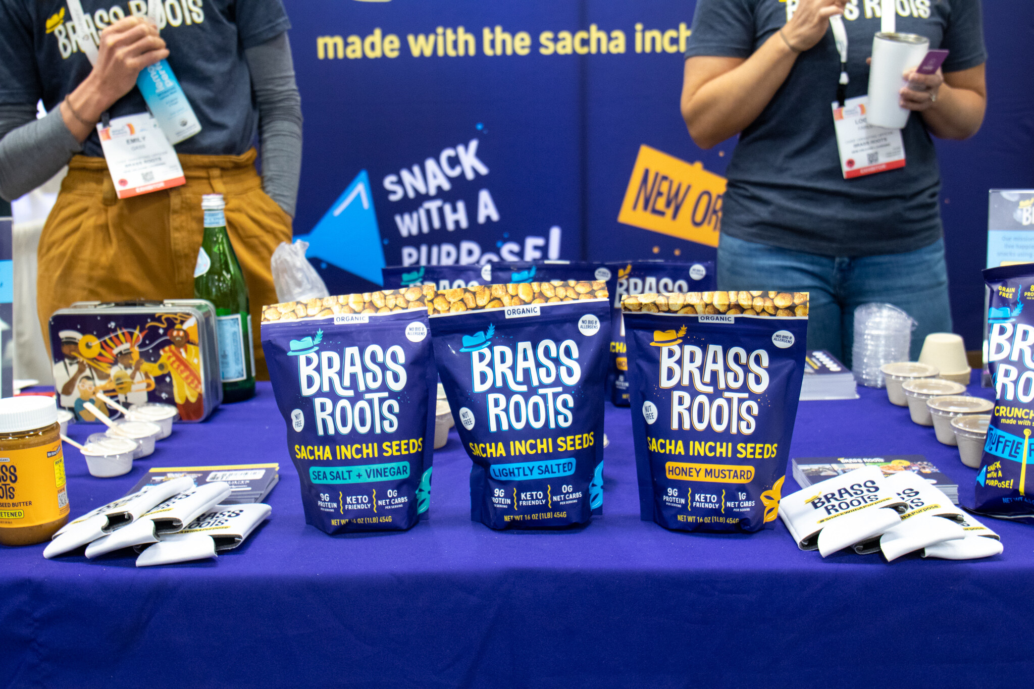 Bags of Brass Roots sacha inchi seeds sit on a purple table with sample cups while staff stand behind
