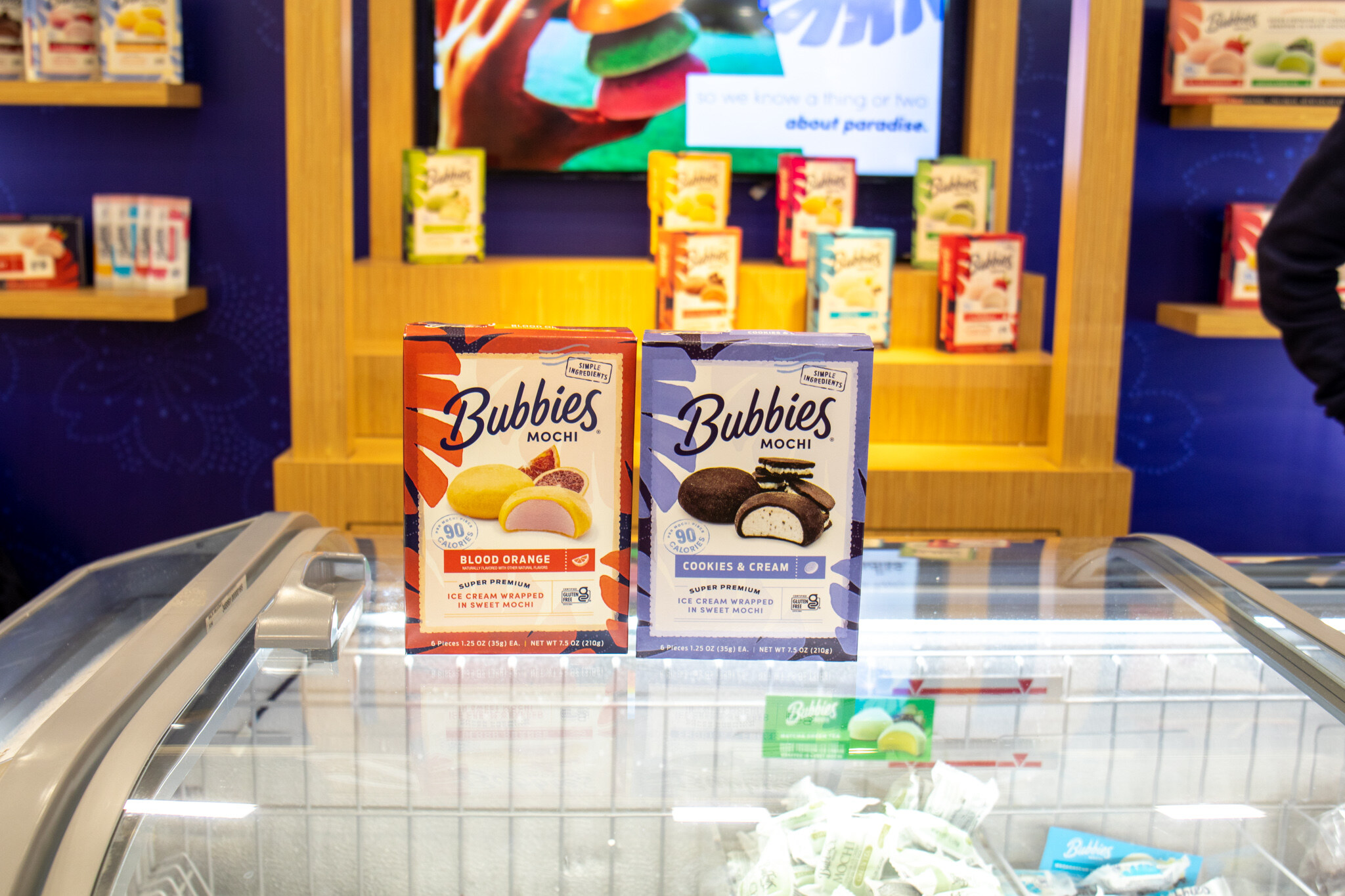 Two boxes of Bubbies mochi ice cream stand on a glass freezer case in front of a blue and wood booth