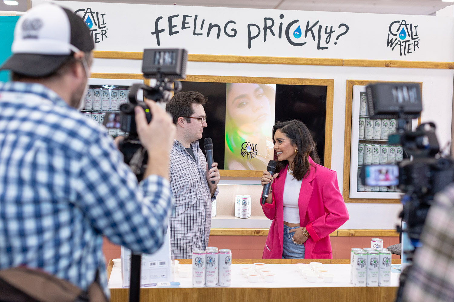 A camera operator films an interviewer and a woman in a pink blazer in front of a Feeling Prickly sign for Cali Water