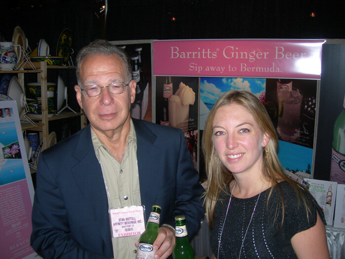 Stan Rottell of Affinity Beverage Inc, at left in glasses and a blue blazer, holds Barritts Ginger Beer bottles beside a smiling blonde woman in front of a Barritts Ginger Beer sign.
