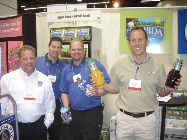 Four men with badges pose in the Espana Brands / Marquis Brands booth. Two hold bottles of water, one holds orange juice and one a dark drink. Lambda and Mondariz signs show.