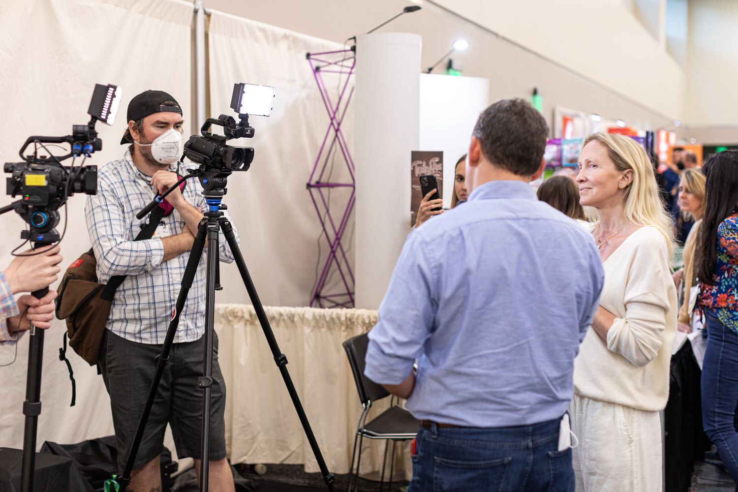 Two camera operators on tripods film while a man in a blue shirt and a woman in white stand nearby