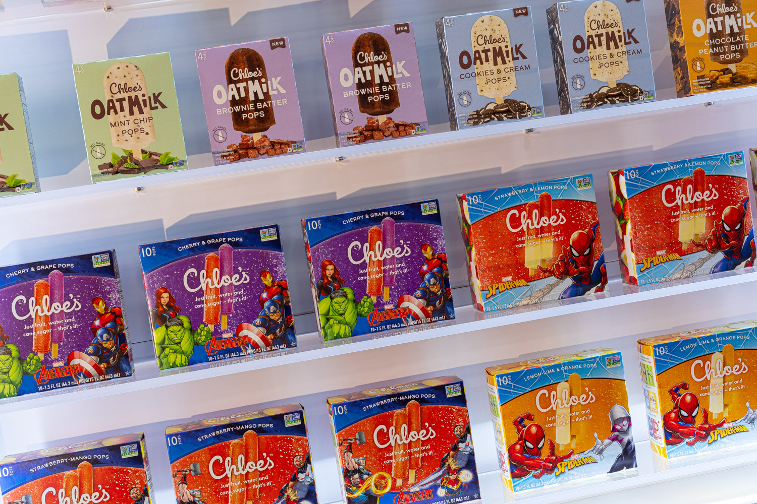 Boxes of Chloe's oat milk and fruit pops, some with Marvel character art, are arranged on white wall shelves