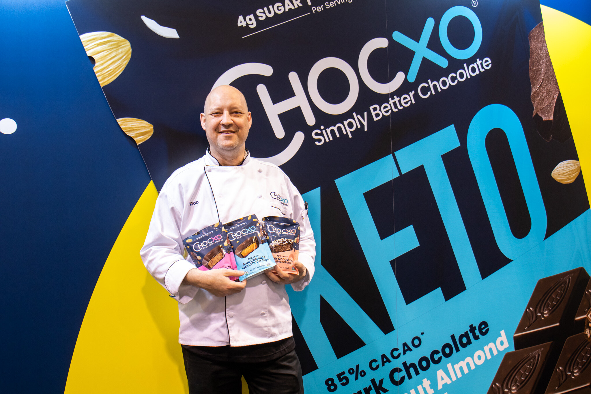 A smiling man in a white chef coat holds Chocxo chocolate packages in front of a Chocxo Keto backdrop