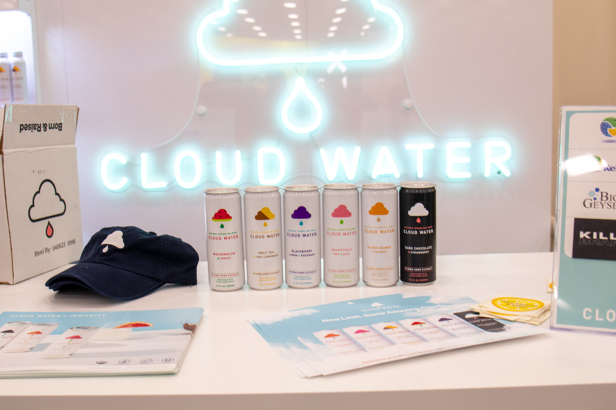 A neon Cloud Water sign glows above a row of Cloud Water cans, a navy hat and brochures on a white counter