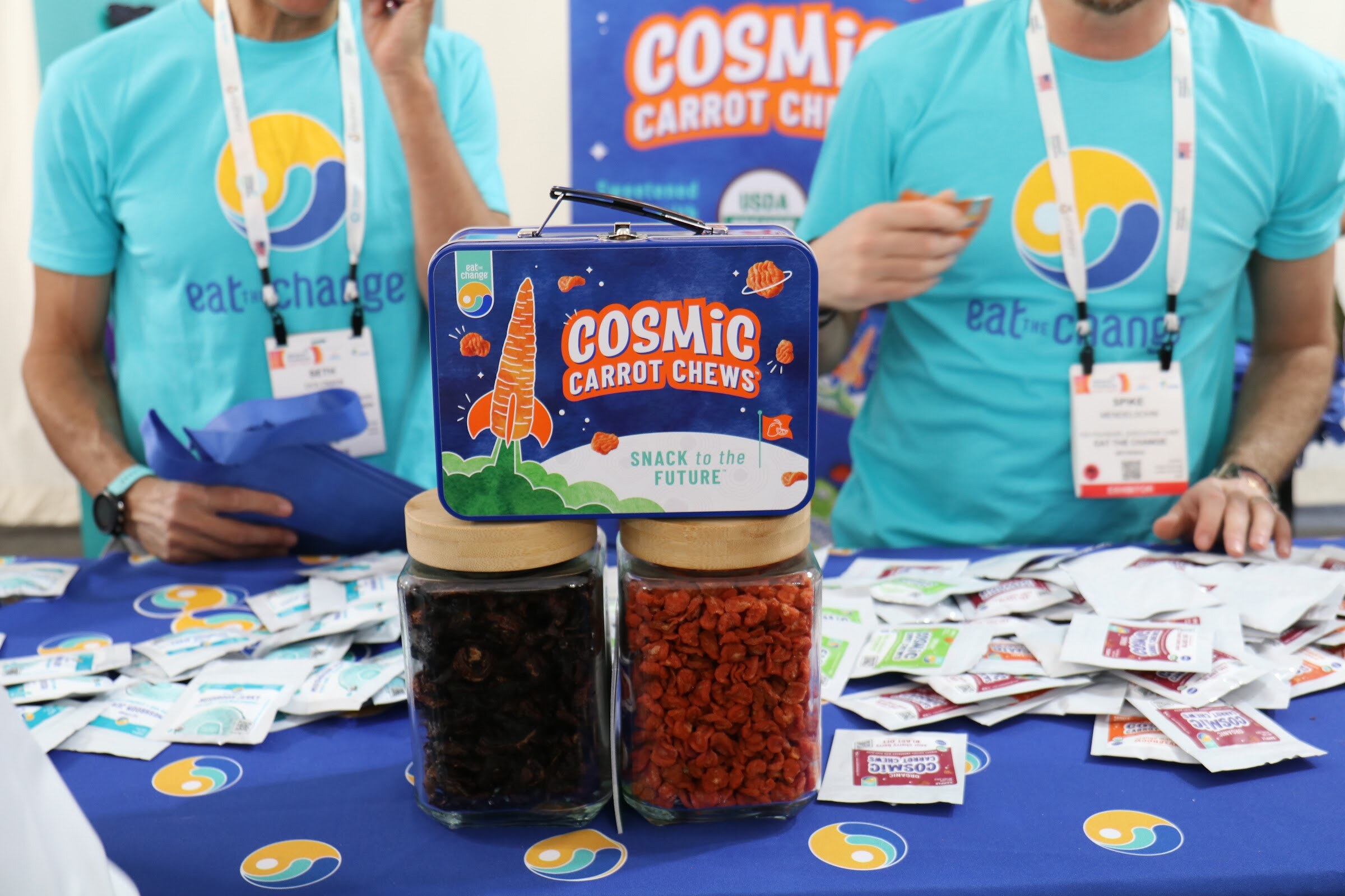 A Cosmic Carrot Chews lunchbox sits on top of jars of snacks on a blue table scattered with samples as booth staff in teal shirts work behind