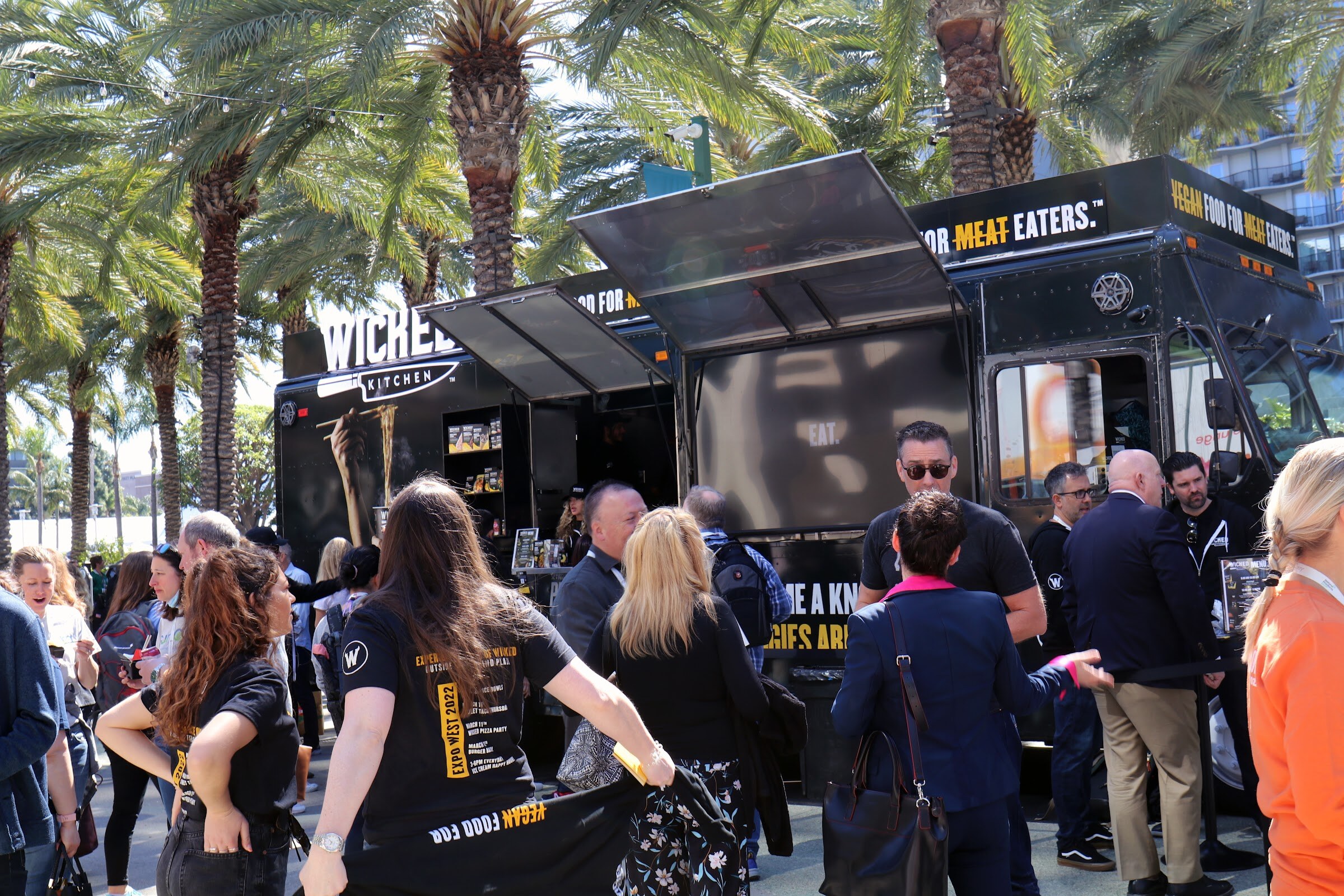 Attendees gather around a black Wicked Kitchen food truck with its serving window open among palm trees