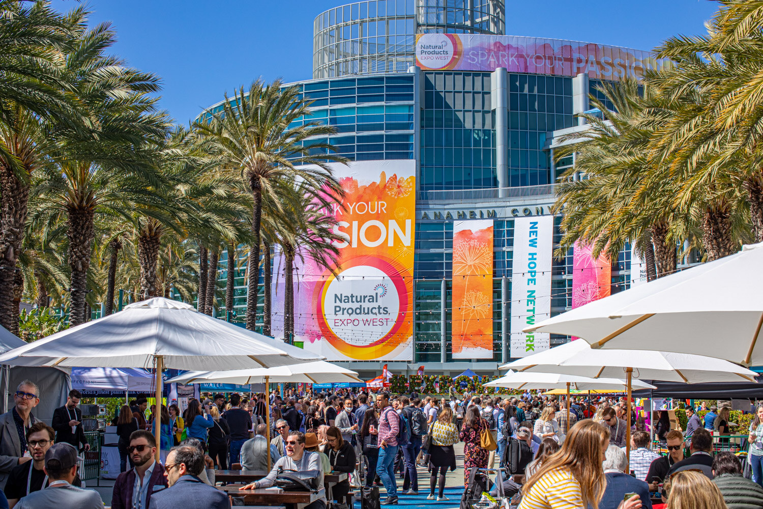 Attendees gather at picnic tables and umbrellas on the plaza in front of the glass Anaheim Convention Center hung with large Natural Products Expo West banners