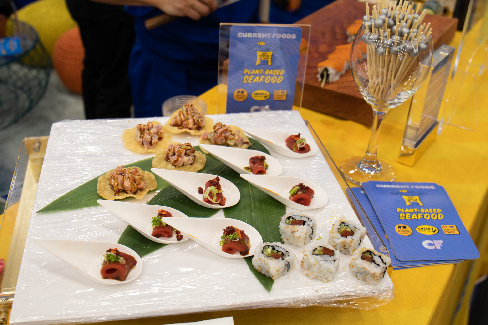 A platter of plant-based tuna bites, sushi rolls and tostadas sits in front of a Current Foods plant-based seafood sign