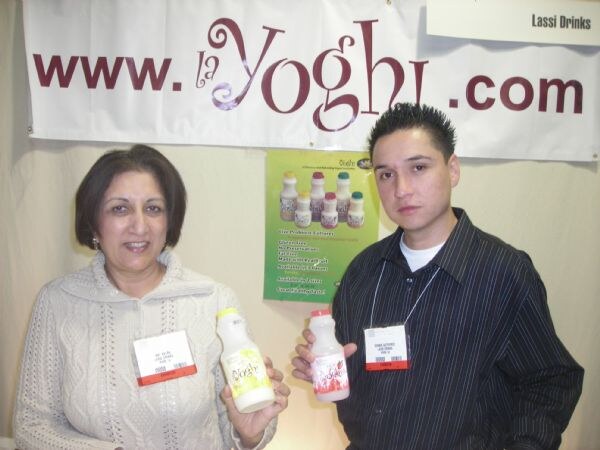 A woman in a white knit sweater and a man in a dark striped shirt, both wearing badges, hold bottles of lassi drinks under a banner reading www.layoghi.com.
