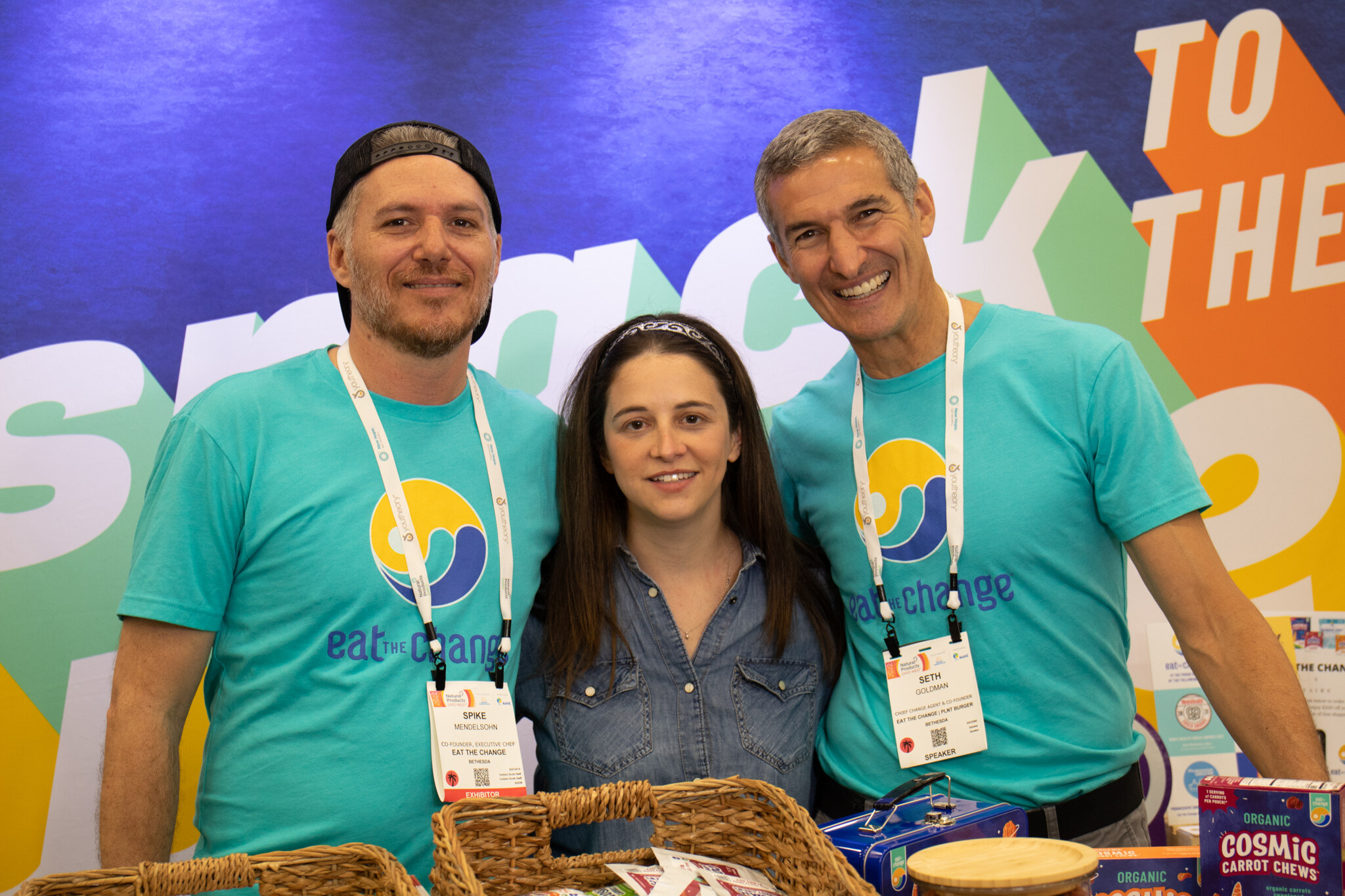 Two men in teal Eat the Change shirts and a woman in a denim shirt smile together behind a basket of snacks at the booth