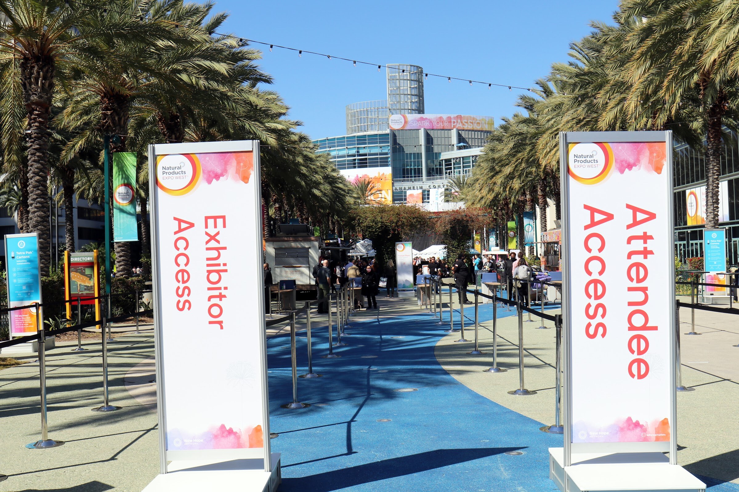 Two tall signs reading Exhibitor Access and Attendee Access stand on a blue walkway lined with palm trees leading toward the convention center