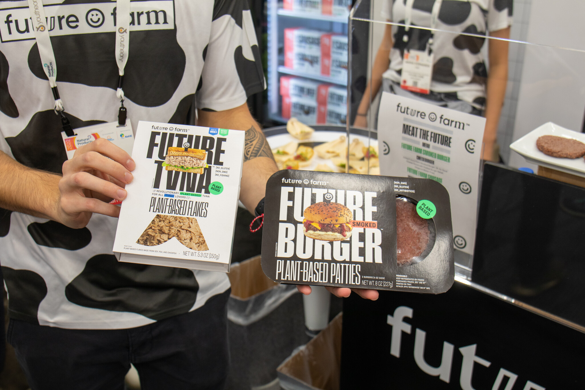 A person in a cow-print shirt holds a Future Tuna package and a Future Burger tray in front of the Future Farm sign