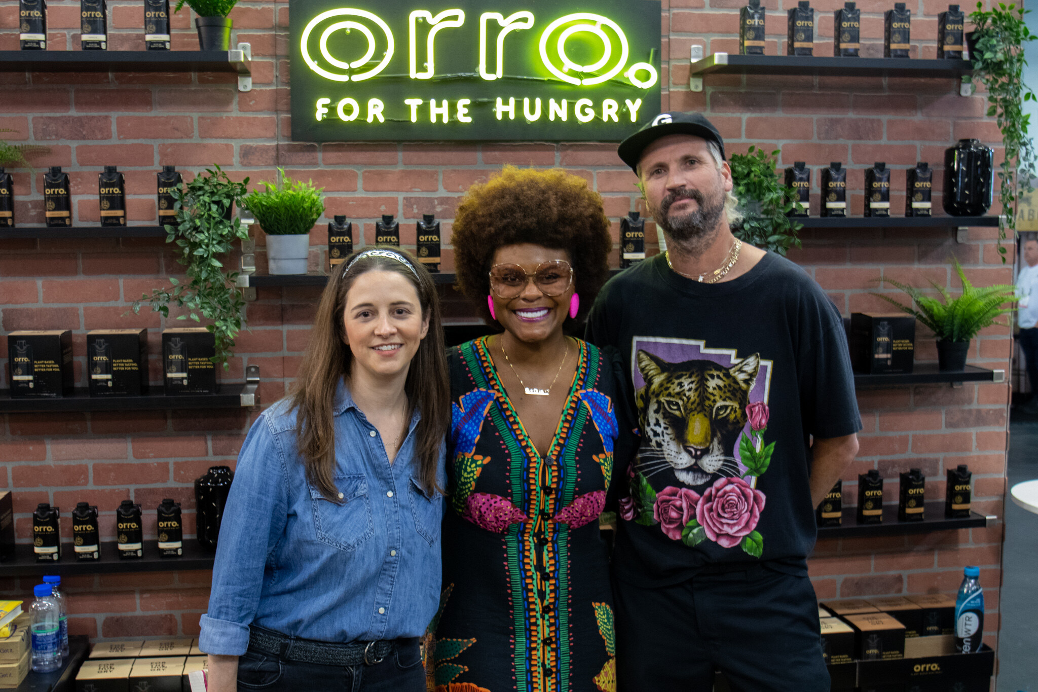 Three people smile together in front of a brick wall with a lit Orro For the Hungry neon sign and shelves of products