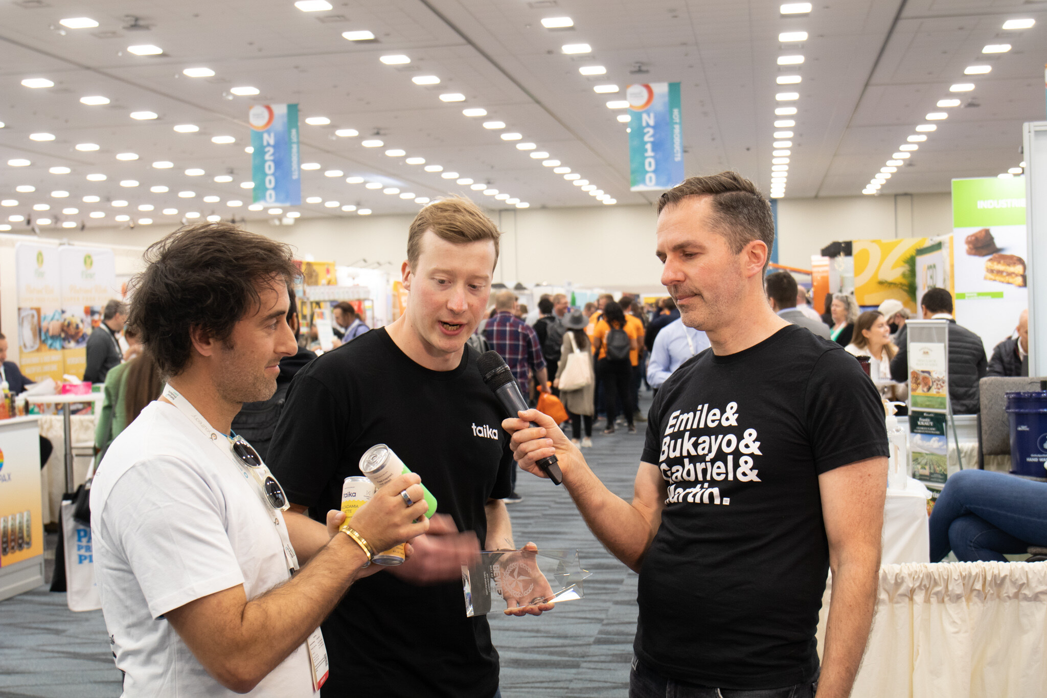 A man in a black t-shirt holds a microphone toward two men, one holding a can and a small trophy, in a busy exhibit hall
