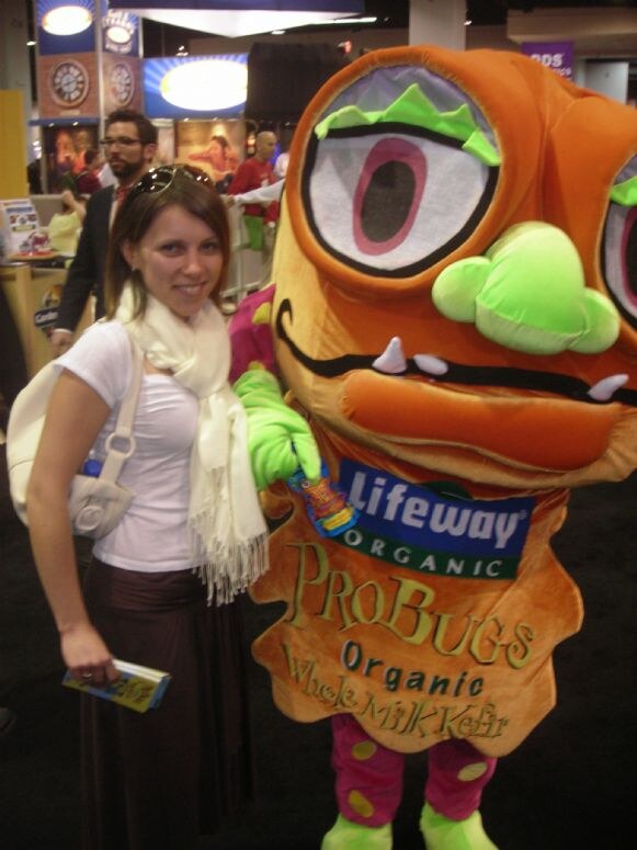 A smiling woman with a white scarf stands beside an orange ProBugs mascot costume with a large eye and a Lifeway Organic logo on its front.