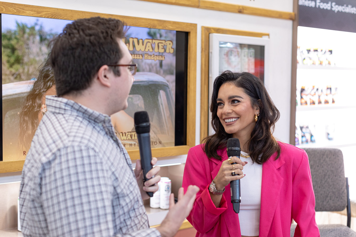 A man holding a microphone interviews a smiling woman in a bright pink blazer at a booth with Cali Water cans on the counter