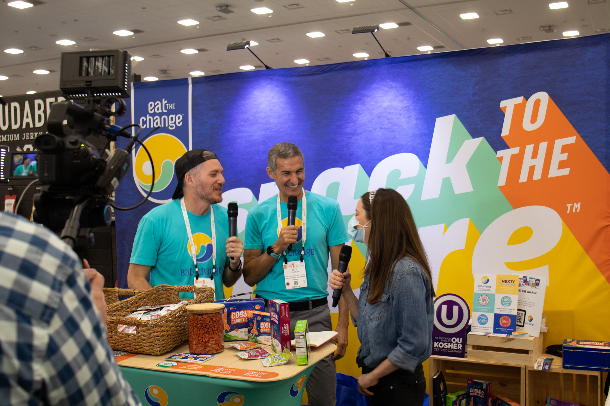Two men in teal Eat the Change shirts and a woman hold microphones at a booth with a blue Snack to the Future backdrop while a camera records