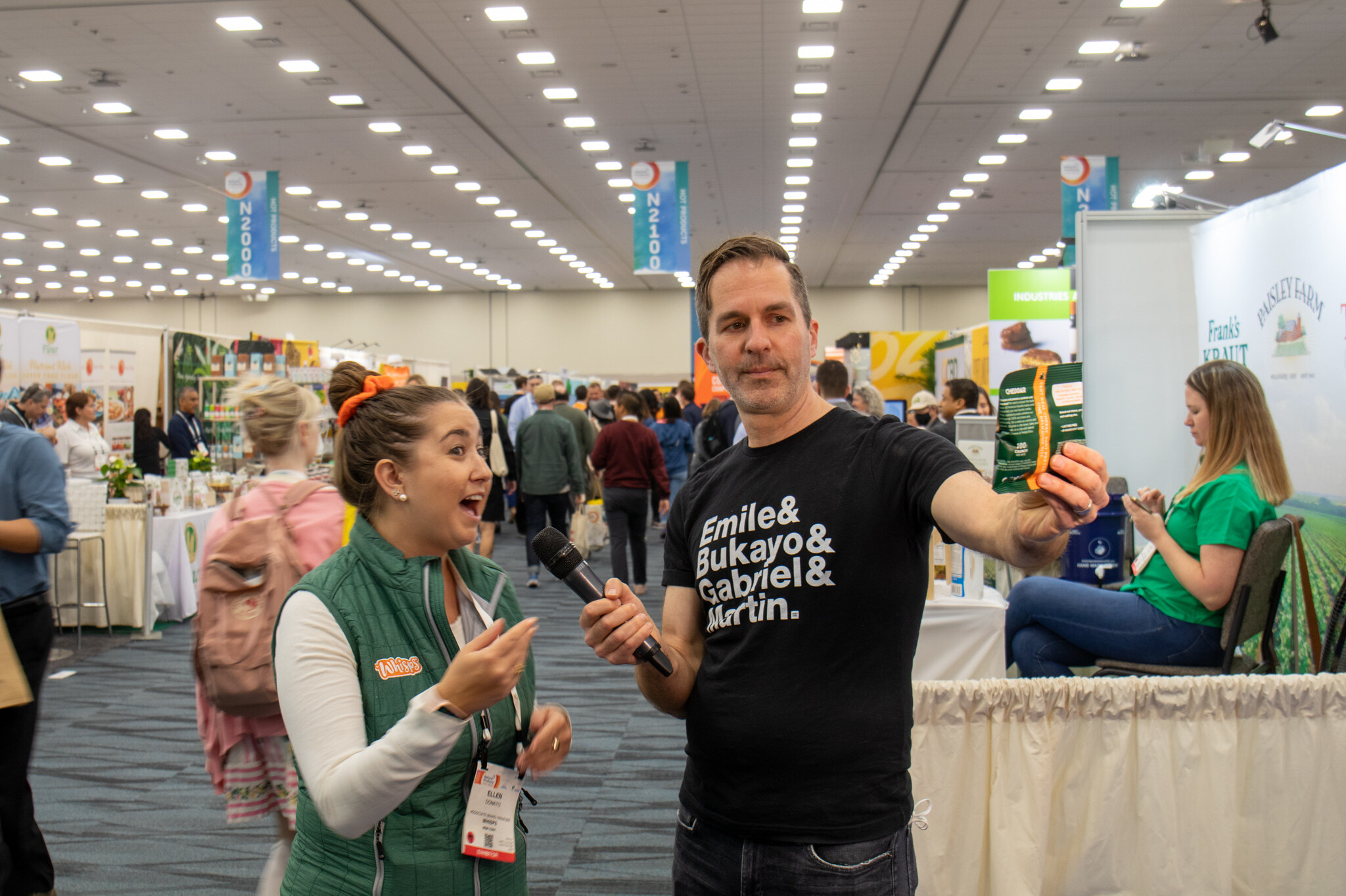 A man in a black shirt holds up a product and a microphone while a woman in a green vest reacts in the exhibit hall