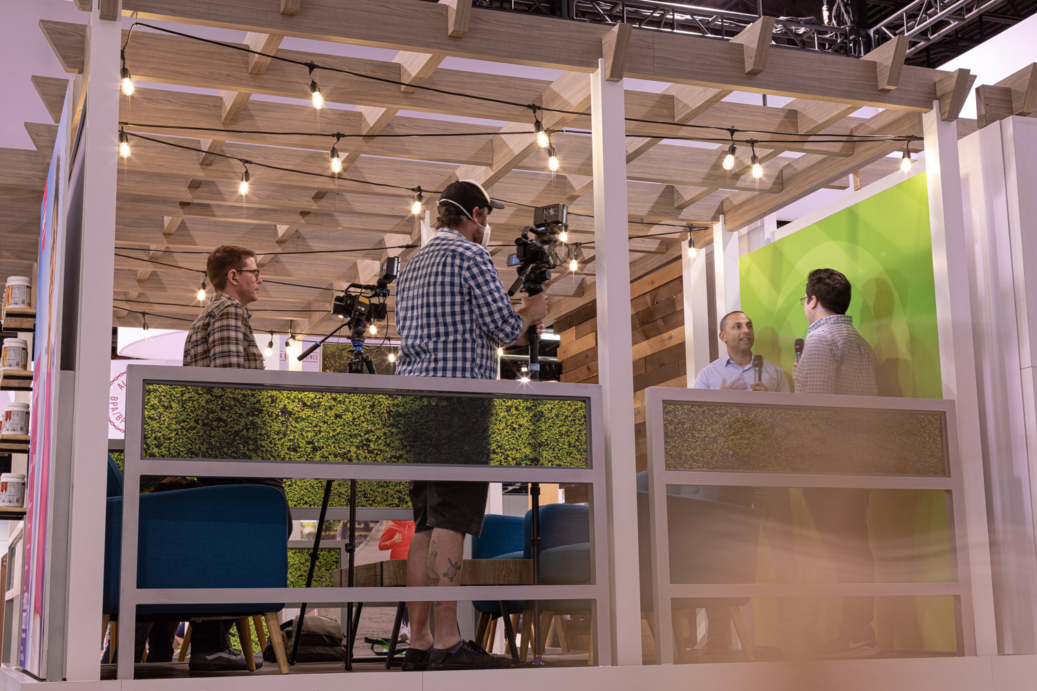 A camera operator films an interview on a raised pergola-style booth stage with string lights and a green backdrop