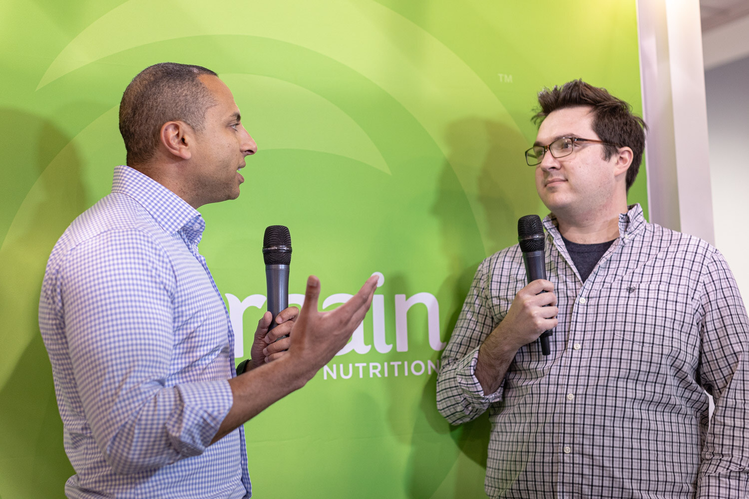 Two men hold microphones and talk in front of a bright green branded backdrop during a video interview