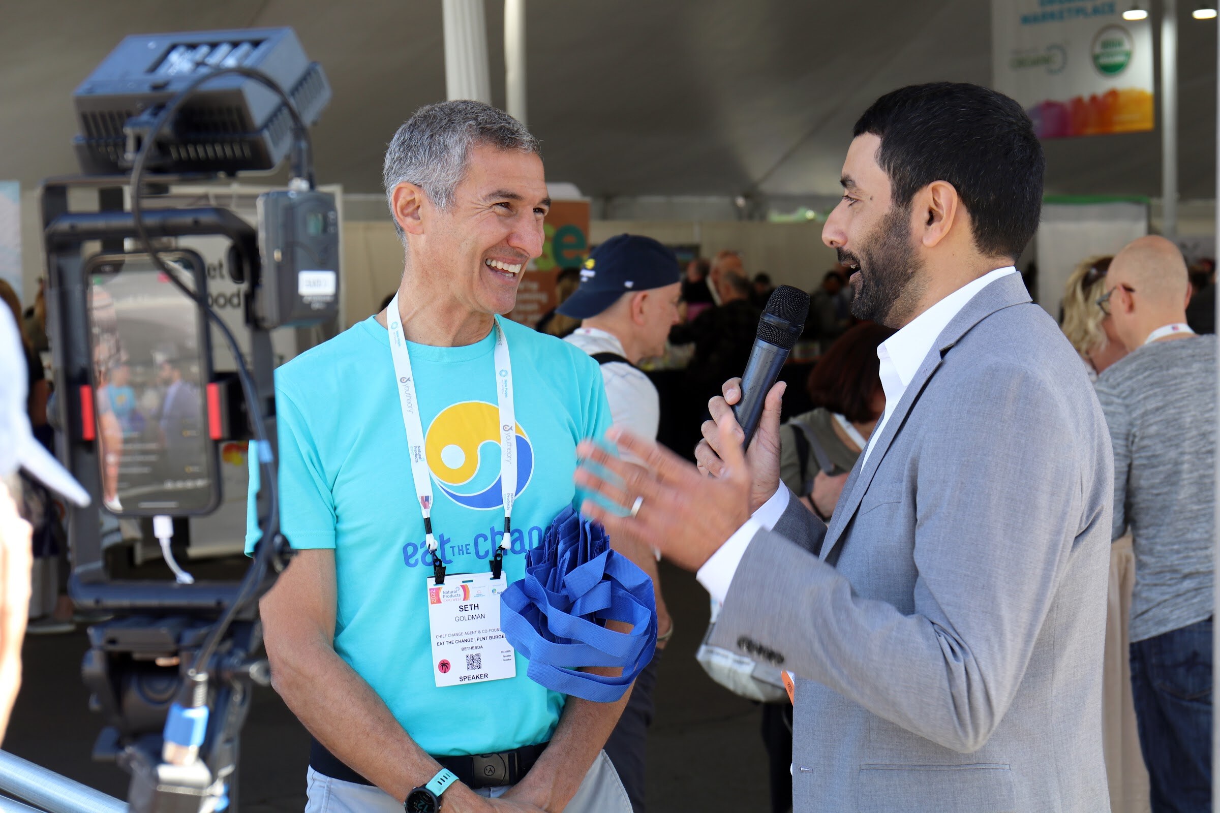 A smiling man in an Eat the Change shirt talks with an interviewer holding a microphone beside a phone livestream rig under a tent