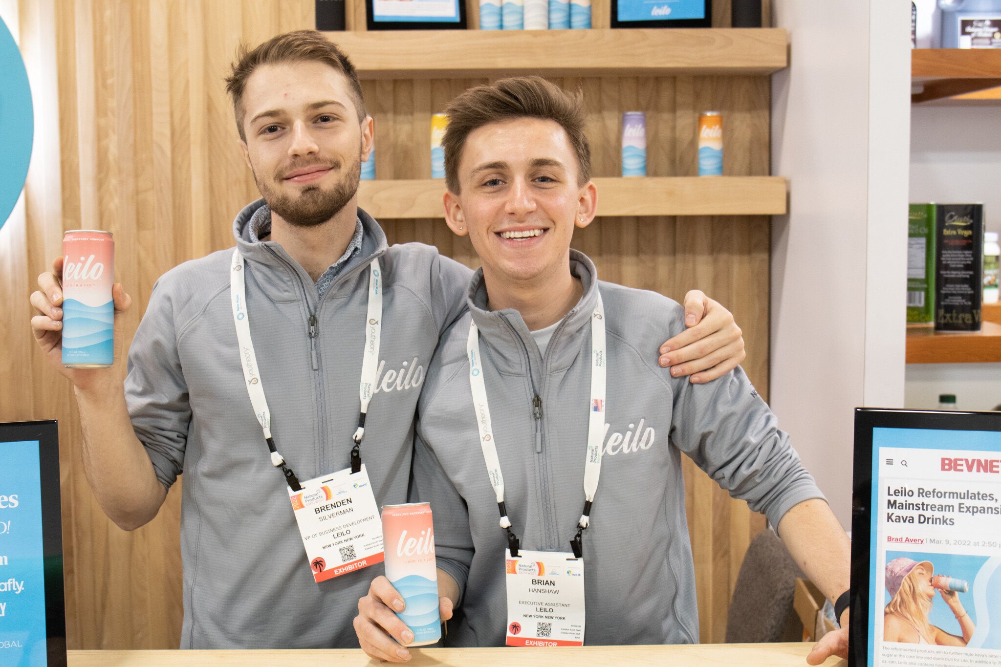 Two smiling men in grey Leilo pullovers hold cans of Leilo at the booth counter