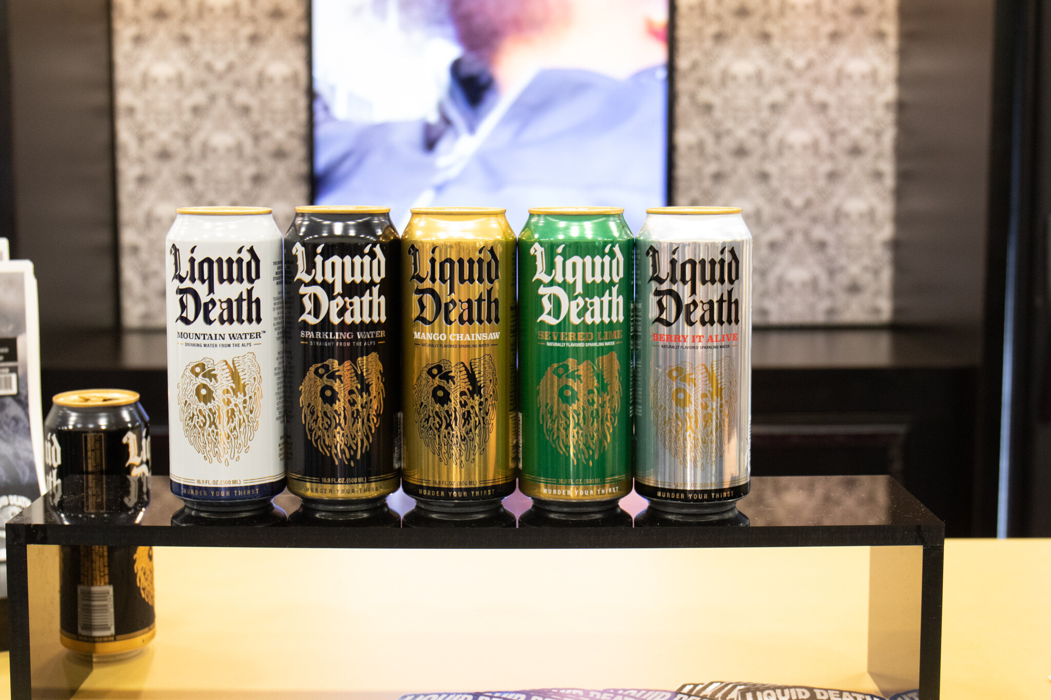 Five Liquid Death cans in white, black, gold, green and silver stand in a row on an illuminated shelf