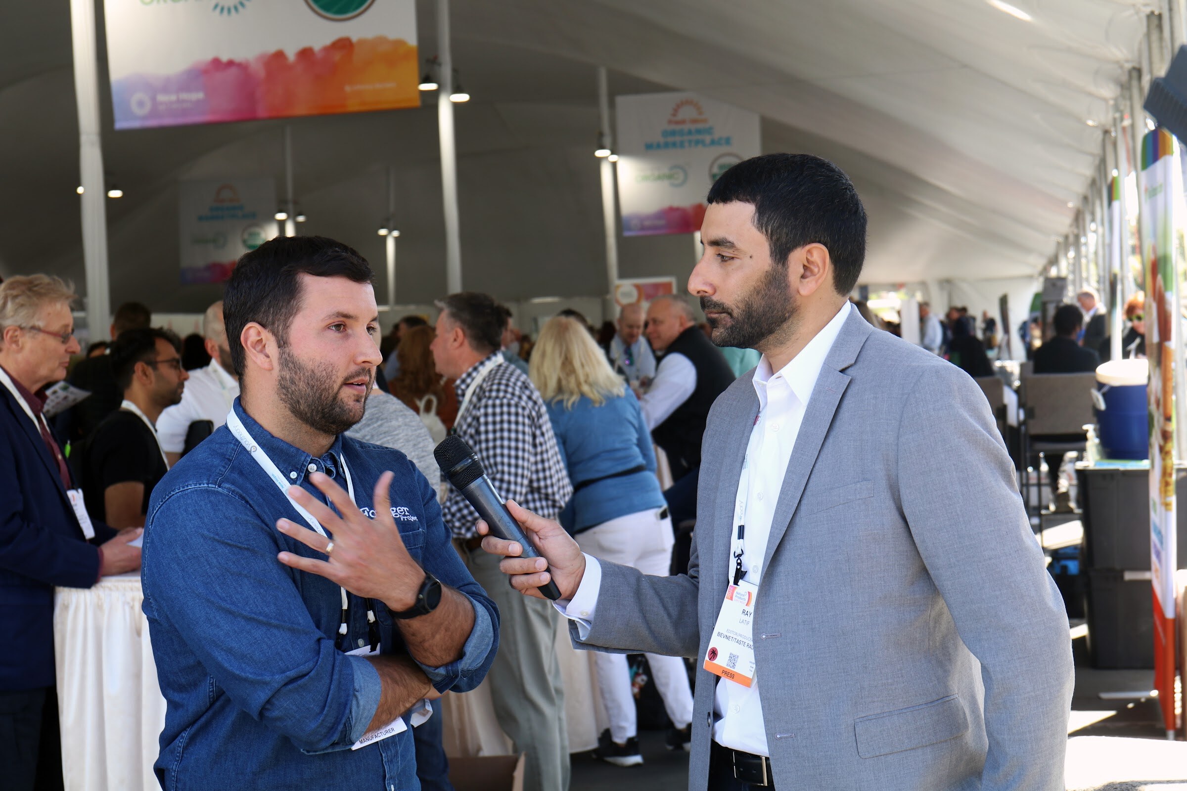 A man in a blue work shirt answers questions from an interviewer with a microphone in a busy tent, with a phone rig recording
