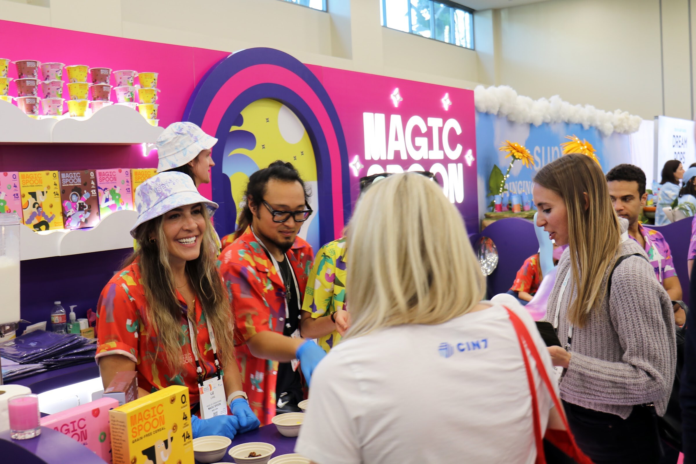 Staff in bright floral shirts and gloves serve samples to visitors at the Magic Spoon booth under a rainbow-shaped sign